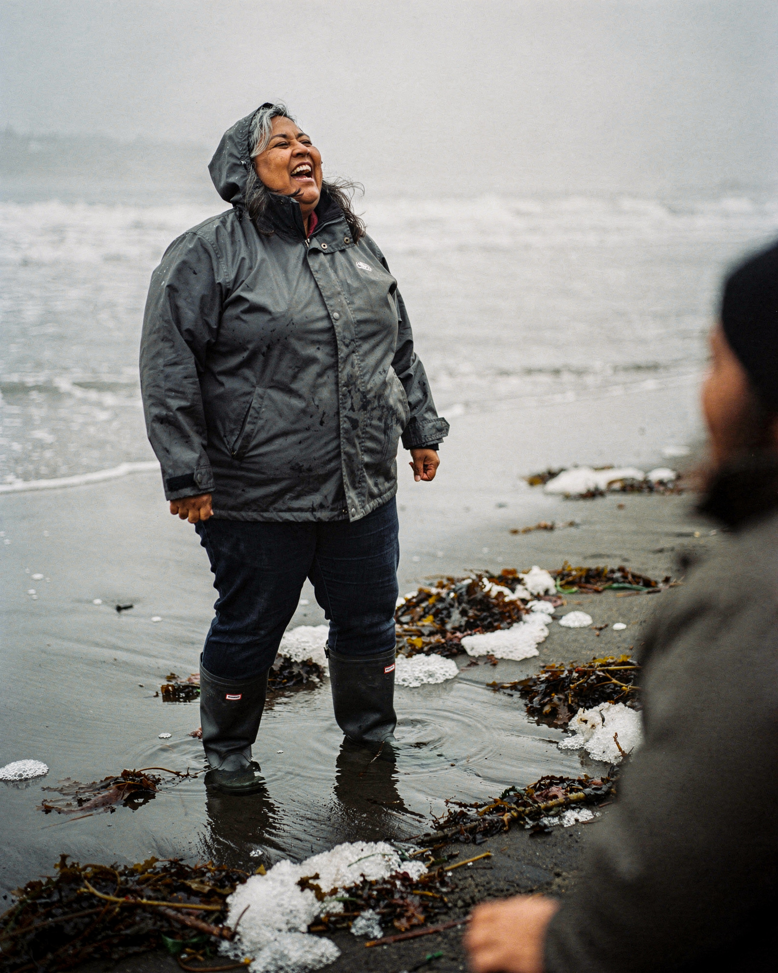 A woman in a rain jacket laughing on a grey beach